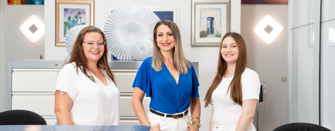 Empfang 1 Das Empfangsteam vom Zahnarzt Dachau-Ost steht lächelnd hinter dem Empfangstresen: Saskia, Seher und Nudzejma.