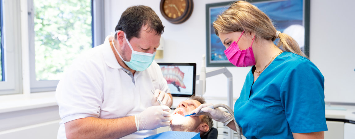 Der Dachauer Zahnarzt Dr. Gitt und Assistentin behandeln einen Patienten auf dem blauen Behandlungsstuhl.